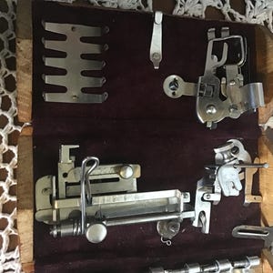 May include: A wooden storage box with a dark maroon velvet lining containing various silver-toned metal sewing machine attachments, including a presser foot, a metal guide plate, and several specialized mechanical components for fabric stitching.