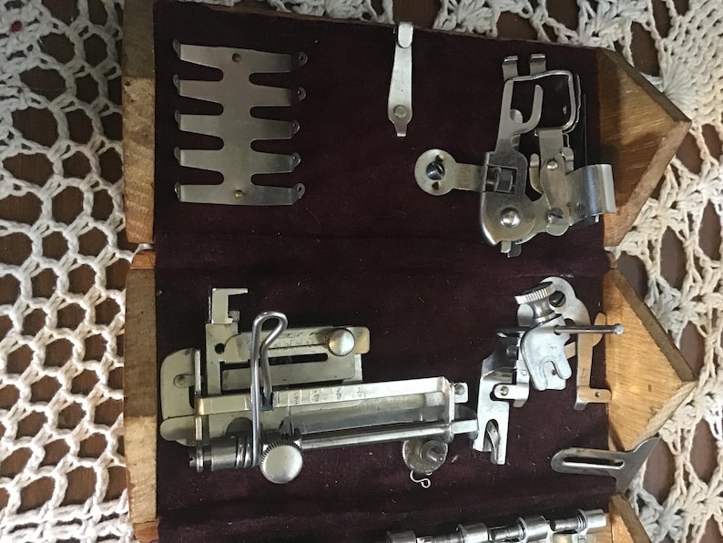 May include: A wooden storage box with a dark maroon velvet lining containing various silver-toned metal sewing machine attachments, including a presser foot, a metal guide plate, and several specialized mechanical components for fabric stitching.