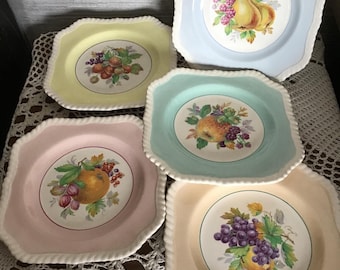 Puede incluir: Conjunto de cinco platos cuadrados de cerámica en colores pastel: amarillo, azul, verde, rosa y beige. Cada plato presenta un diseño central de frutas, incluyendo peras, uvas y naranjas, rodeado por un borde blanco en forma de cuerda.
