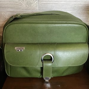 May include: A vintage olive green camera bag with a shoulder strap. The bag features a front flap with a silver-toned metal clasp and a small pocket. The text "Camera MD" is visible on the front. The bag is made of a textured material.
