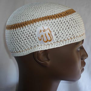 May include: White and gold kufi hat with an embroidered patch featuring Arabic calligraphy. The hat has a woven design with a gold band around the crown. The hat is displayed on a mannequin head.