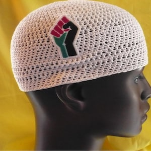 May include: A white crocheted skull cap with a patch featuring a raised fist in red, green, and black. The cap is made of a loosely woven material, and the patch is on the left side. The background is a solid yellow.