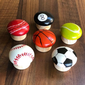 May include: Six painted wooden knobs with different sports designs. The knobs are round and have a white base. The designs include a baseball, basketball, football, tennis ball, 8-ball, and a red and white striped ball.