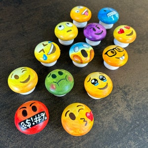Op de afbeelding: Twaalf ronde houten ornamenten geschilderd met verschillende gele, blauwe, groene, rode en zwarte emoji-gezichten. De gezichten omvatten een knipperend gezicht, een huilend gezicht, een verbaasd gezicht, een gezicht dat zijn tong uitsteekt, een gezicht met hartjesogen, een verward gezicht, een glimlachend gezicht, een gezicht dat pruilt, een gezicht met een bedekte mond, een gezicht met een verband, een gezicht met een snor en een gezicht met een kroon.
