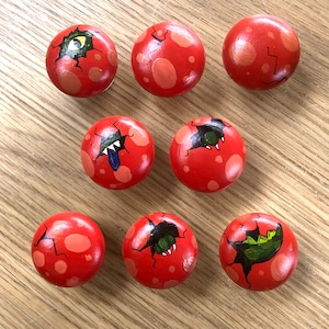 May include: A collection of eight red, round, wooden knobs, each with a unique hand-painted design. Some knobs feature cracked eggshell patterns, while others depict cartoon monster faces with green eyes and open mouths. The knobs are arranged on a light brown wooden surface.