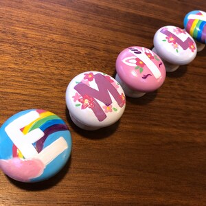 May include: Five colourful round knobs with painted designs. Each knob features a different letter of the alphabet, a rainbow, a unicorn, and flowers.