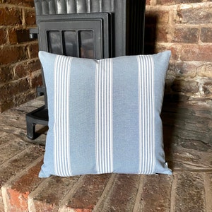 May include: A square, light blue pillow with vertical white stripes. The pillow is made of a soft fabric and is placed on a brick surface. The pillow is a decorative item, suitable for a living room or bedroom.