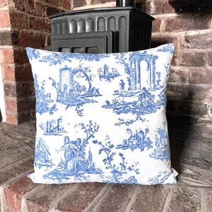 May include: A white throw pillow with a blue toile pattern featuring a repeating design of trees, buildings, and figures. The pillow is sitting on a brick surface.