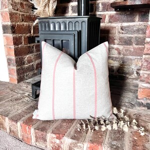 May include: A grey and white striped cushion with red stripes. The cushion is sitting on a brick surface in front of a fireplace.