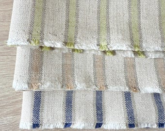 May include: Close-up of three folded linen fabric samples with vertical stripes. The top sample has green stripes, the middle has tan stripes, and the bottom has blue stripes. Each sample has a frayed edge.