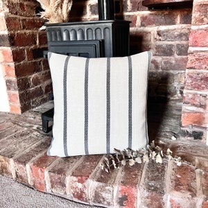 May include: A white and black striped throw pillow with a subtle texture. The pillow is sitting on a brick surface in front of a black wood-burning stove.