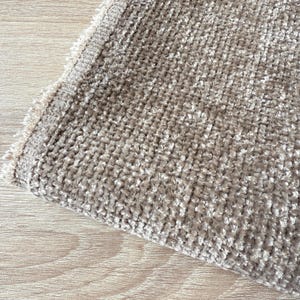 May include: A close-up shot of a beige fabric with a textured, woven design. The material has a soft, plush feel and a fringed edge. The fabric is folded, showing the texture and construction of the textile.