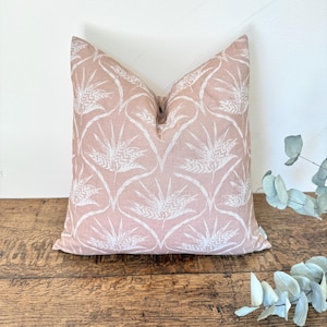 May include: A square cushion with a pale pink backdrop and a white wheat design. The cushion is positioned on a wooden surface. This is a home decor item.