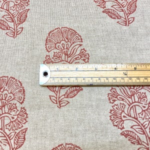 May include: Close-up of a beige fabric with a repeating pattern of rust-coloured floral designs. A ruler is placed on the fabric to show the scale of the pattern.