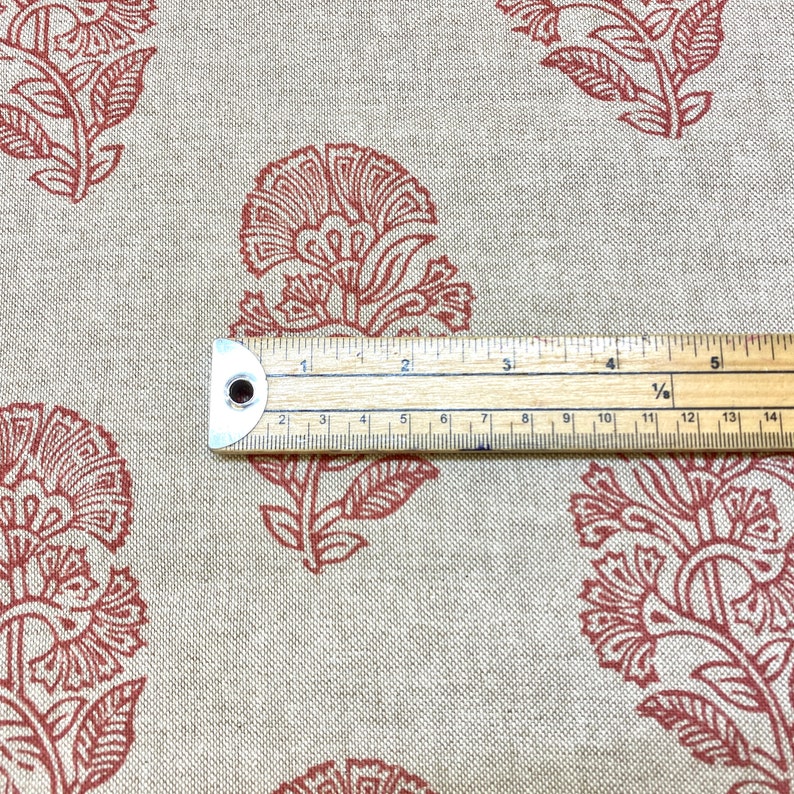 May include: Close-up of a beige fabric with a repeating pattern of rust-coloured floral designs. A ruler is placed on the fabric to show the scale of the pattern.