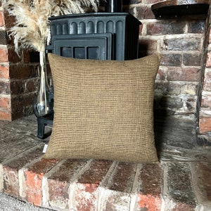 May include: A brown and beige square cushion with a subtle textured pattern. The cushion is sitting on a brick hearth in front of a black wood-burning stove.