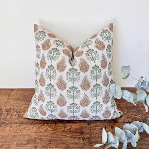 Puede incluir: Un cojín decorativo cuadrado con un patrón repetido de diseños de cachemira y florales en tonos marrón, verde y blanco. El cojín está colocado sobre una superficie de madera.