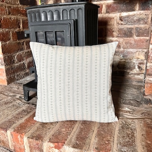 May include: A cream-coloured throw pillow with a subtle black and white floral pattern. The pillow is sitting on a brick surface in front of a black wood-burning stove.