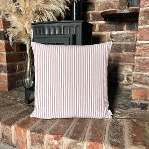 May include: Square pillow with a white background and vertical red stripes. The pillow is placed on a brick surface in front of a black stove. The pillow has a tag on the bottom right corner.