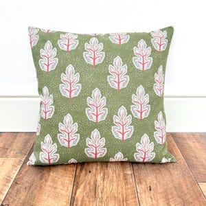 May include: Square cushion featuring a repeating pattern of white and red leaf designs on a green background. The cushion is placed on a wooden surface.
