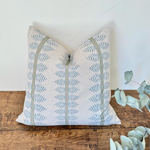 May include: Square cushion with a light beige background and a repeating pattern of blue leaf-like shapes. Vertical stripes of light green border the leaf pattern. The cushion is placed on a wooden surface.
