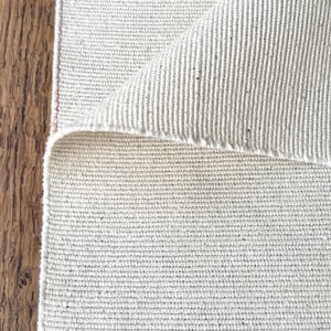 May include: A rectangular off-white fabric rug with a visible woven texture and a folded corner revealing the underside. The material features a consistent grid-like weave pattern throughout, showcasing a neutral, light-coloured textile surface.