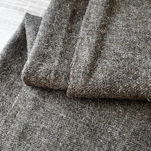 以下が含まれることがあります： 濃い灰色のツイード生地の折り畳まれた部分のクローズアップ。生地は、より明るい色の斑点のあるテクスチャ加工された表面を持っています。折り畳まれた部分の端が見え、素材の厚さと織り方を示しています。