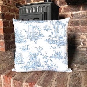 Può includere: Un cuscino bianco con un motivo toile blu che presenta un design ripetitivo di alberi, edifici e figure. Il cuscino è appoggiato su una superficie di mattoni.