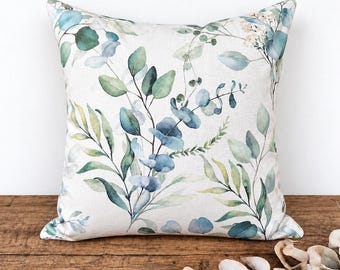 May include: A square throw cushion featuring a watercolour print of eucalyptus branches and leaves in shades of sage green, teal, and blue on a light off-white fabric background. The botanical pattern covers the entire front surface of the cushion.