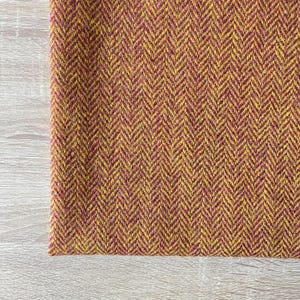 May include: Close-up of a section of fabric with a herringbone pattern. The fabric features a blend of mustard yellow and burgundy colors. The texture appears to be woven, with a slightly rough surface.