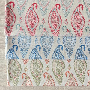 Op de afbeelding: Drie gevouwen stukken stof met een paisley patroon in rood, blauw en groen. Het paisley ontwerp wordt herhaald over de stof, met een neutrale achtergrond. De stoffen zijn op elkaar gestapeld.