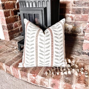 May include: A decorative cushion with a white background and a pattern of green leaves and brown stripes. The cushion is sitting on a brick surface.