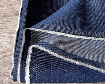 Op de afbeelding: Close-up van gevouwen donkerblauwe denimstof met witte gerafelde randen. De stof is getextureerd en lijkt nieuw te zijn. De afbeelding toont de details van de weefsel en de kleur van de denim.