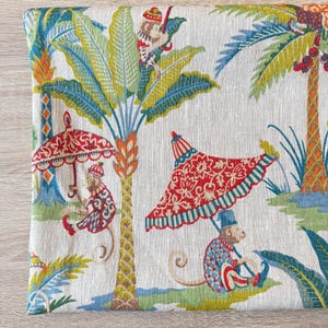 Puede incluir: Una tela estampada con un diseño tropical caprichoso. El patrón presenta monos, palmeras y sombrillas decorativas en tonos rojos, azules, verdes y amarillos sobre un fondo blanco. La tela tiene una textura similar al lino.