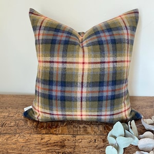 Puede incluir: Un cojín decorativo cuadrado con un estampado de cuadros en tonos verdes, azules, rojos y crema. El cojín está hecho de una tela texturizada y se encuentra sobre una superficie de madera. Es un artículo de decoración para el hogar.