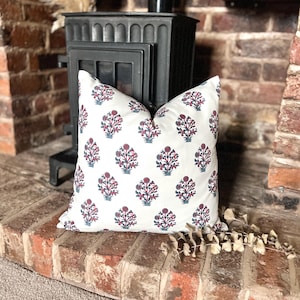 May include: A white cushion with a red and blue floral pattern. The cushion is sitting on a brick surface in front of a black wood-burning stove.