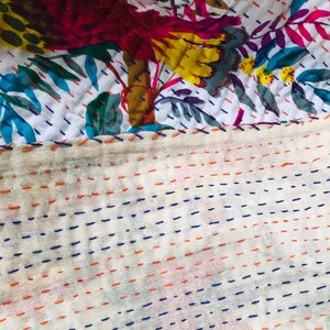 May include: Close-up of a white fabric with a floral pattern and a stitched border. The fabric is stitched with blue and orange thread in a straight line pattern.
