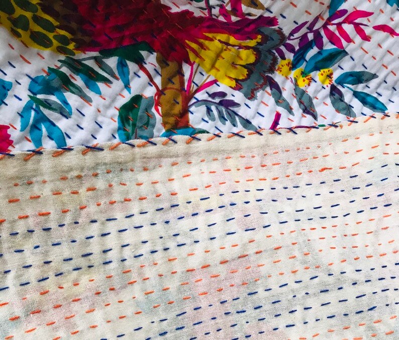 May include: Close-up of a white fabric with a floral pattern and a stitched border. The fabric is stitched with blue and orange thread in a straight line pattern.