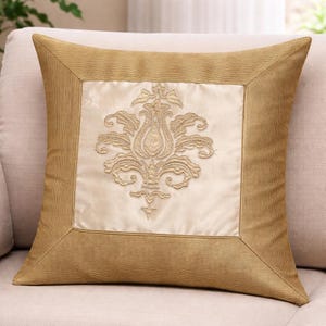 Pode incluir: Almofada decorativa quadrada com uma borda dourada e um painel central com um desenho floral bordado em ouro. A almofada é de cor bege neutra e parece ser feita de um tecido macio.