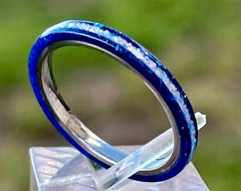 Op de afbeelding: Een dunne blauwe ring met witte opaal glitter ingebed in de band. De ring wordt getoond op een heldere acryl standaard.