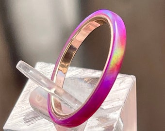 May include: A thin, iridescent pink ring with a gold band. The ring is on a clear display stand.