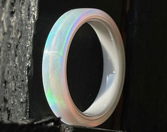 May include: A white opal ring with iridescent colors. The ring has a smooth, polished finish and a minimalist design. The opal displays flashes of green, blue, and pink. The ring is set against a dark, textured background.