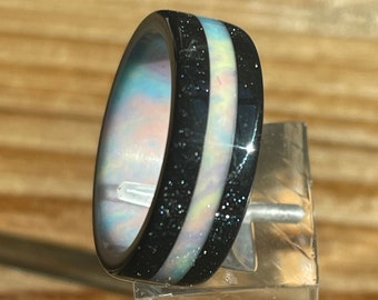 May include: A black and white ring with a sparkly iridescent inlay. The ring is on a clear display stand.