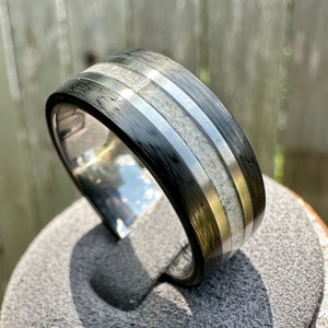 May include: A black and silver ring with a textured finish and two white stripes. The ring is on a gray display stand.