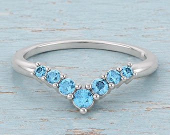 May include: A silver-toned chevron ring featuring seven round-cut light blue stones set along the V-shaped band. The center stone is the largest, with three smaller stones graduating in size on each side, creating a symmetrical design.