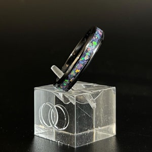 May include: A black ring with a band of iridescent opal inlay. The ring is displayed on a clear acrylic ring stand.