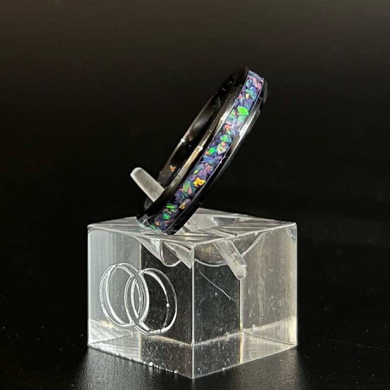 May include: A black ring with a band of iridescent opal inlay. The ring is displayed on a clear acrylic ring stand.