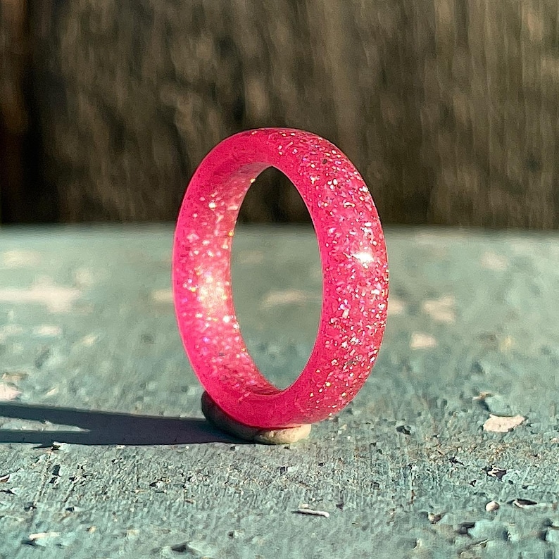 K&ouml;nnte beinhalten: Ein leuchtend pinkfarbener Ring mit eingearbeiteten Glitzerpartikeln. Der Ring ist auf einem kleinen, hellfarbigen Stein platziert, mit einem verwitterten blauen Untergrund. Die Farbe des Rings ist ein helles, auff&auml;lliges Rosa.
