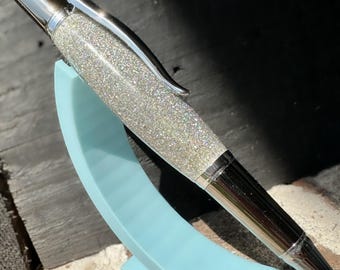 May include: A silver pen with a sparkly, iridescent glitter finish. The pen is on a light blue stand.