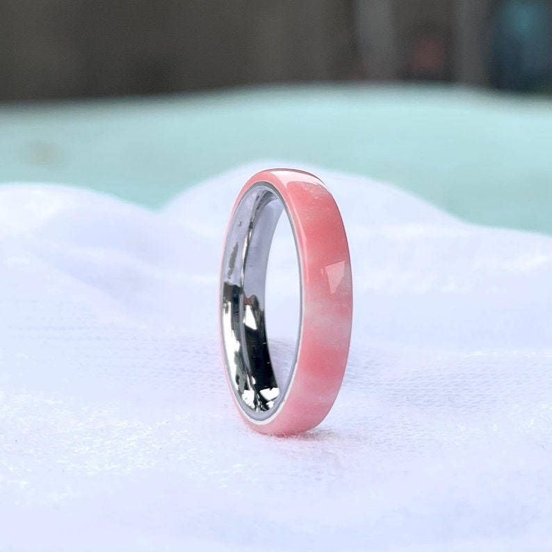K&ouml;nnte beinhalten: Nahaufnahme eines Rings mit einem rosa Band und einem silbernen Innenraum. Der Ring ist vor einem wei&szlig;en Hintergrund platziert. Das rosa Band hat ein marmoriertes Aussehen.
